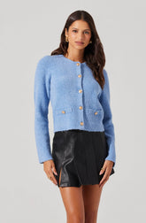 A woman is wearing a light blue textured cardigan with long sleeves and gold buttons down the front. The cardigan has a round neckline and two small pockets at the front. She pairs it with a high-waisted black leather mini skirt with front pleats and side slits.